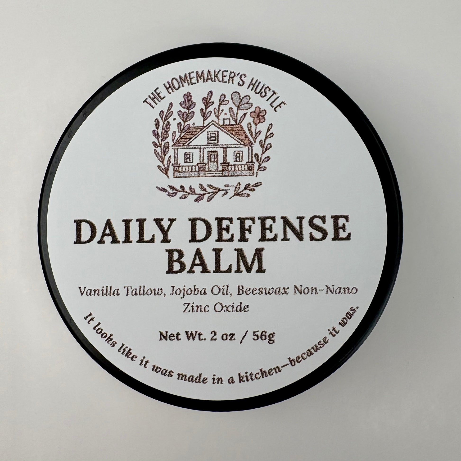Round product label for The Homemaker’s Hustle Daily Defense Balm. The label features an illustrated farmhouse surrounded by branches. Text reads: ‘Daily Defense Balm. Vanilla Tallow, Jojoba Oil, Beeswax, Non-Nano Zinc Oxide. Net Wt. 2 oz / 56 g.’ Small text at the bottom says, ‘It looks like it was made in a kitchen—because it was.’