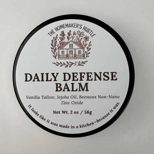 Round product label for The Homemaker’s Hustle Daily Defense Balm. The label features an illustrated farmhouse surrounded by branches. Text reads: ‘Daily Defense Balm. Vanilla Tallow, Jojoba Oil, Beeswax, Non-Nano Zinc Oxide. Net Wt. 2 oz / 56 g.’ Small text at the bottom says, ‘It looks like it was made in a kitchen—because it was.’