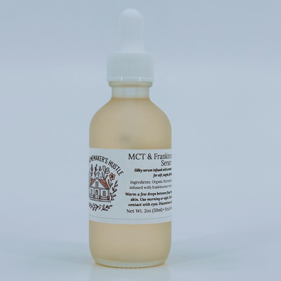 Glass dropper bottle of MCT & Frankincense Serum from The Homemaker’s Hustle. The label includes a farmhouse illustration and text describing a silky serum infused with frankincense resin. Net weight 2 oz.