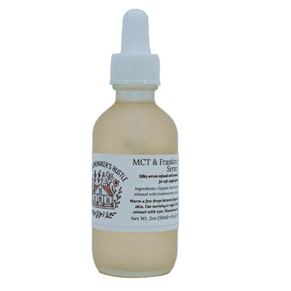 Glass dropper bottle of MCT & Frankincense Serum from The Homemaker’s Hustle. The label includes a farmhouse illustration and text describing a silky serum infused with frankincense resin. Net weight 2 oz.