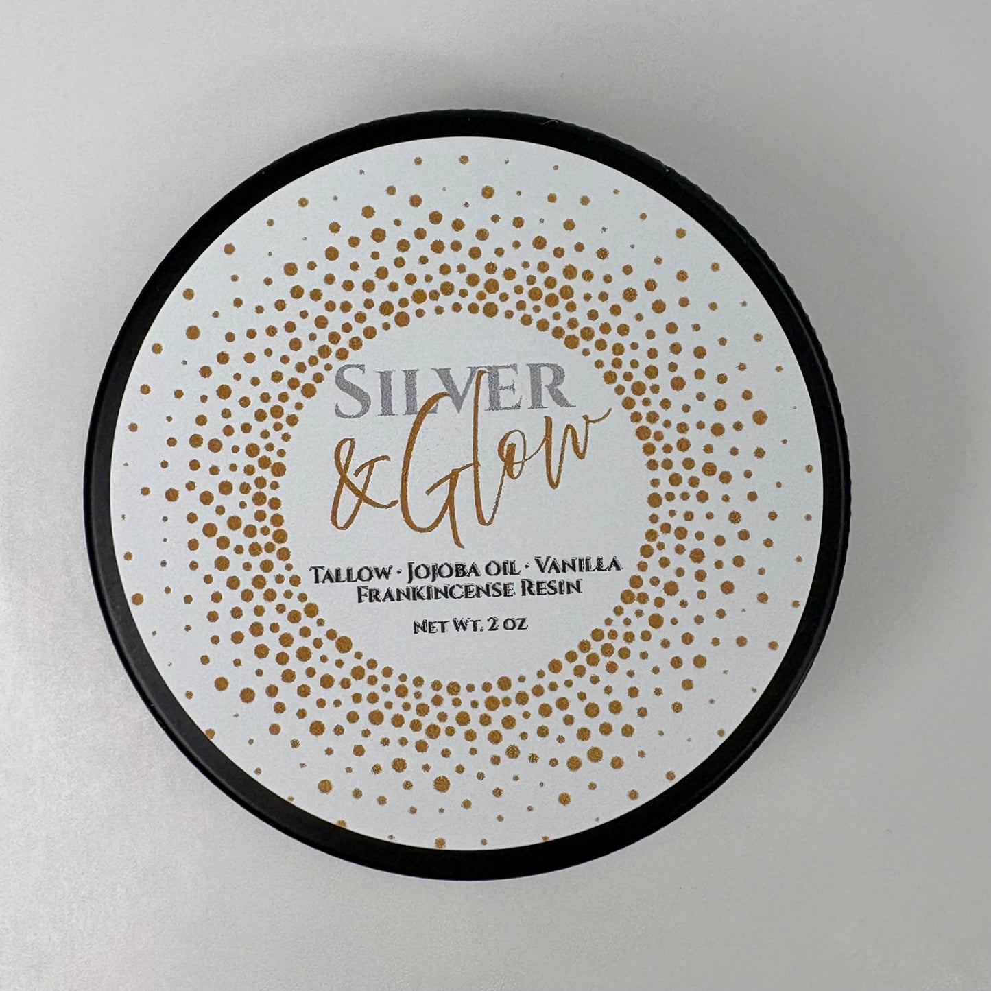Round product label for Silver & Glow balm. The label features a white background with a gold dotted circular pattern. Text reads: ‘Silver & Glow. Tallow, Jojoba Oil, Vanilla, Frankincense Resin. Net Wt. 2 oz.
